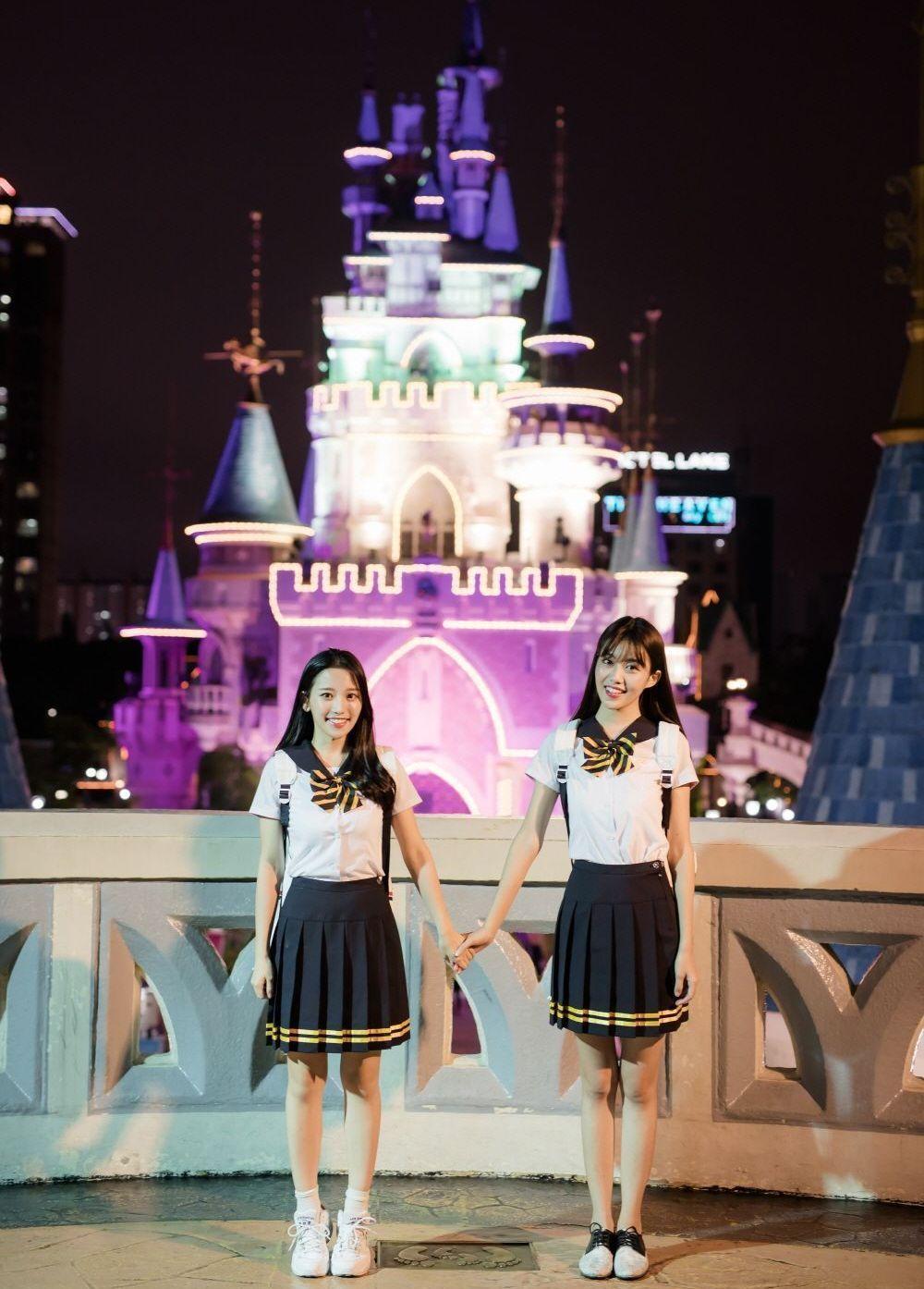 樂天樂園夜景，兩位年輕女孩身穿韓國高中制服，站在樂園的夢幻城堡前拍照，充滿青春活力。