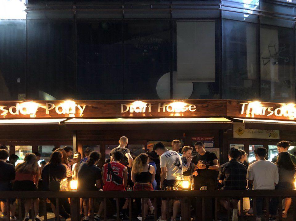 韓國弘大附近的酒吧外觀夜間景象，人群聚集在木質外牆前，燈光和背景音樂營造歡快氛圍。