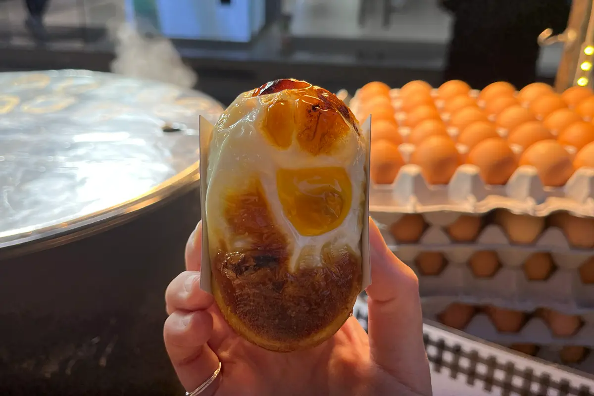 韓國街頭小食雞蛋包，燒烤至金黃酥脆的外皮，手持展示，背景為熱氣騰騰的街邊食攤。