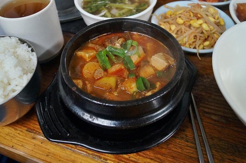 一碗韓式大醬湯，裝在傳統韓國石鍋中，上面浮著豆腐與青蔥，周圍環繞著米飯與韓式小菜，呈現出典型的韓國餐桌。