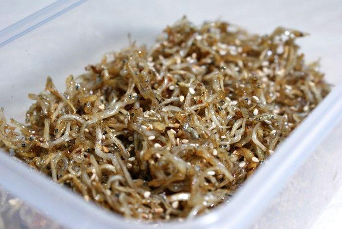 韓式炒小魚乾小菜，裝在透明塑膠盒中，小魚乾細密可見，撒上芝麻與調味料，成為餐桌上的精緻小點。