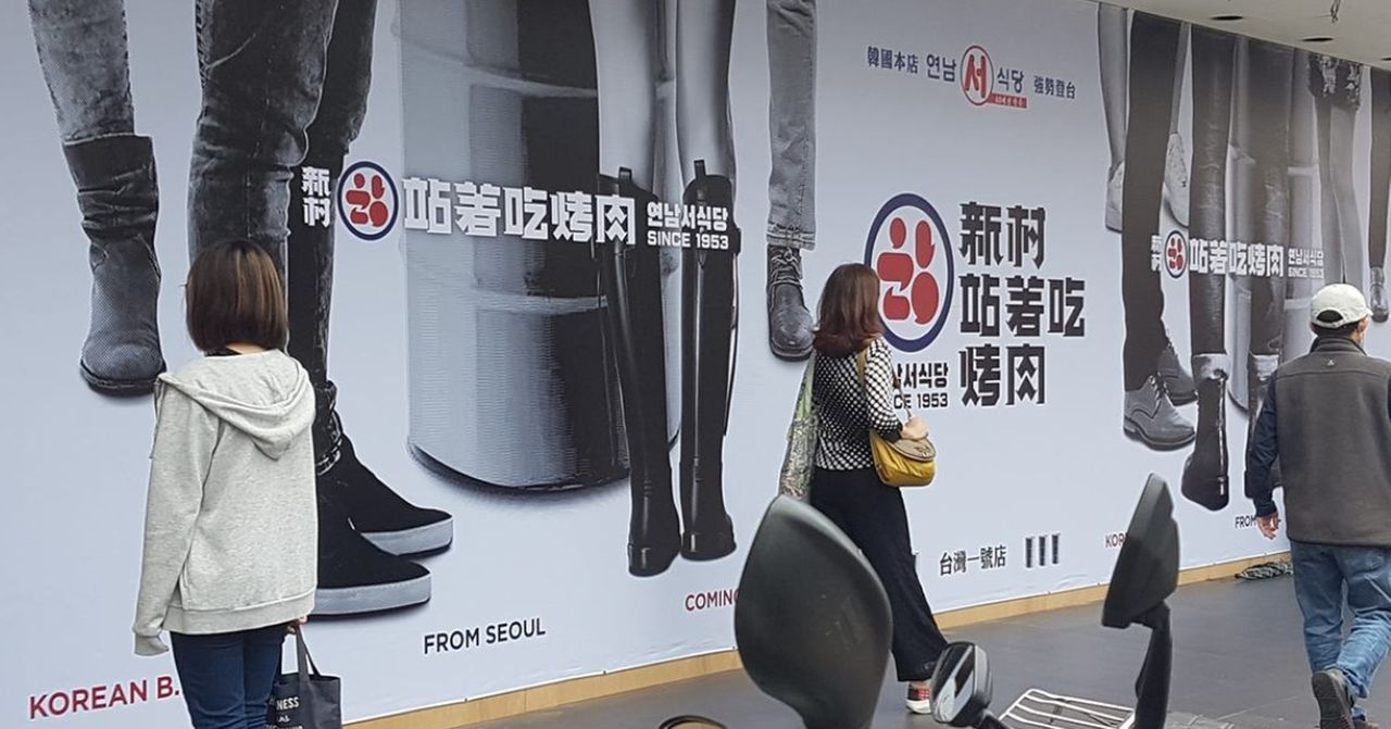 大幅的站著吃烤肉廣告牌，位於街道旁，幾位路人走過，背景呈現臺灣知名品牌的宣傳。