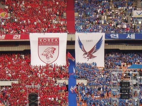 Photo du festival YeonKoJeon entre l'Université de Yonsei et l'Université de Corée avec des supporters en rouge et bleu brandissant des bannières avec les emblèmes universitaires. L'événement de renommée comprend du football, du baseball, du basketball, et plus, attirant une foule nombreuse pour célébrer la rivalité historique entre les deux prestigieuses institutions coréennes.