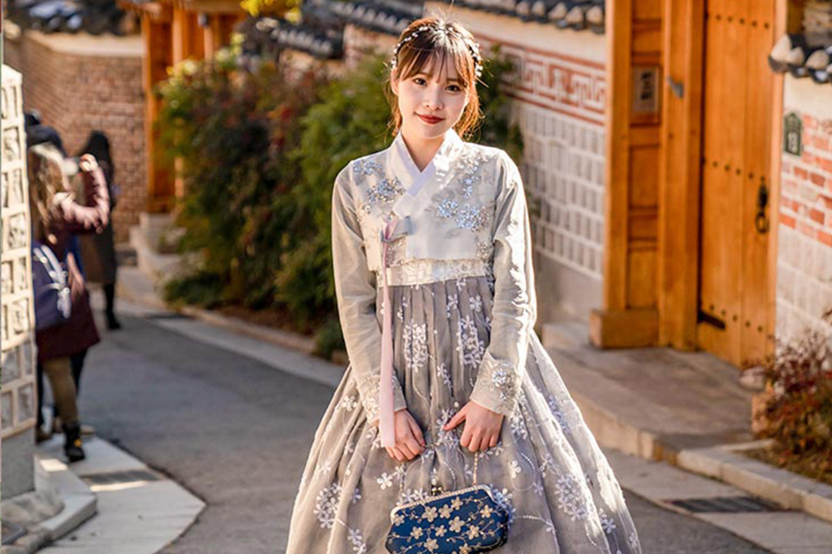 穿著灰色繡花韓服的女子走在傳統韓屋村的小巷中，背景是韓式建築