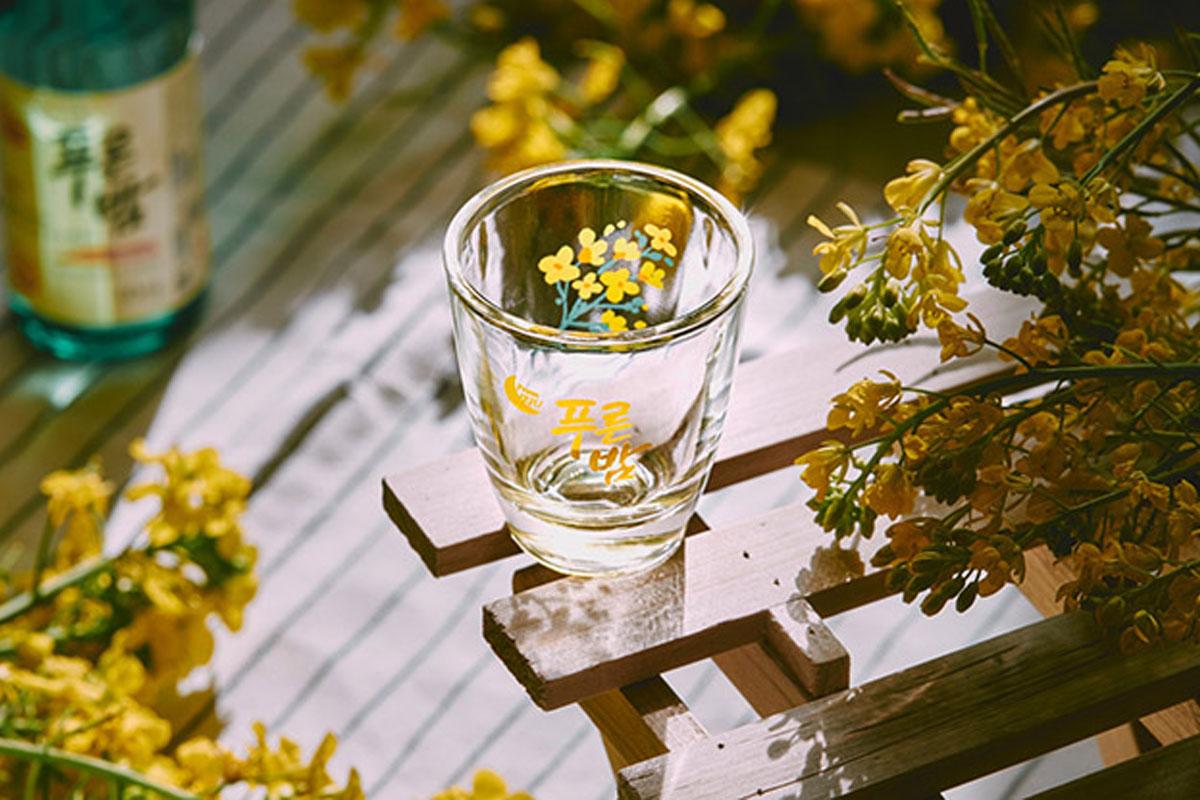 A picture of Jeju soju glass sitting on a wooden table surrounded by flowers with a bottle of soju seen in the distance.