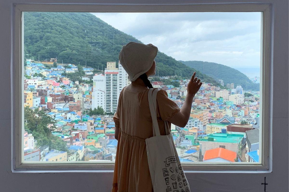 站在窗邊欣賞甘川文化村的女孩，窗外是一片色彩繽紛的房屋和綠意盎然的山丘。