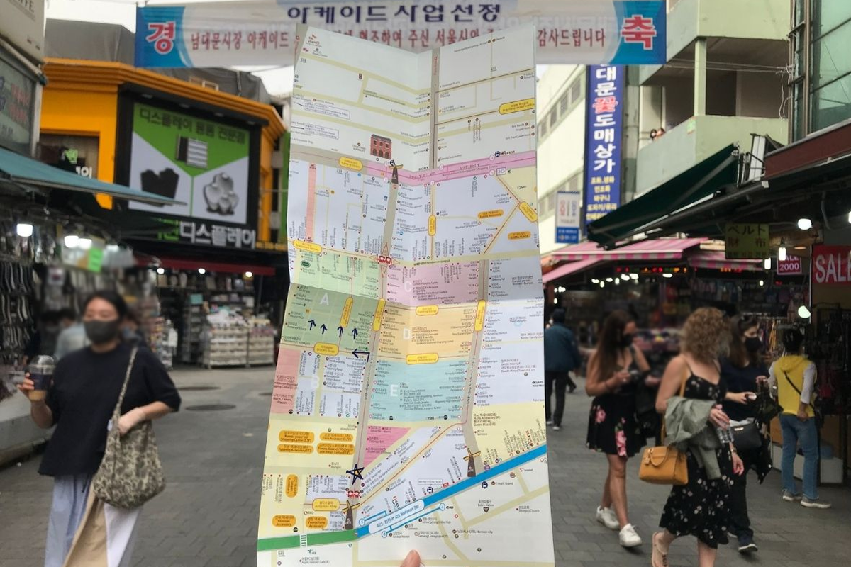 正在探索南大門市場的遊客，手中持有市場地圖，背景是多間開放的店鋪。圖中顯示南大門市場的入口，讓遊客在繁忙的市場中尋找方向。