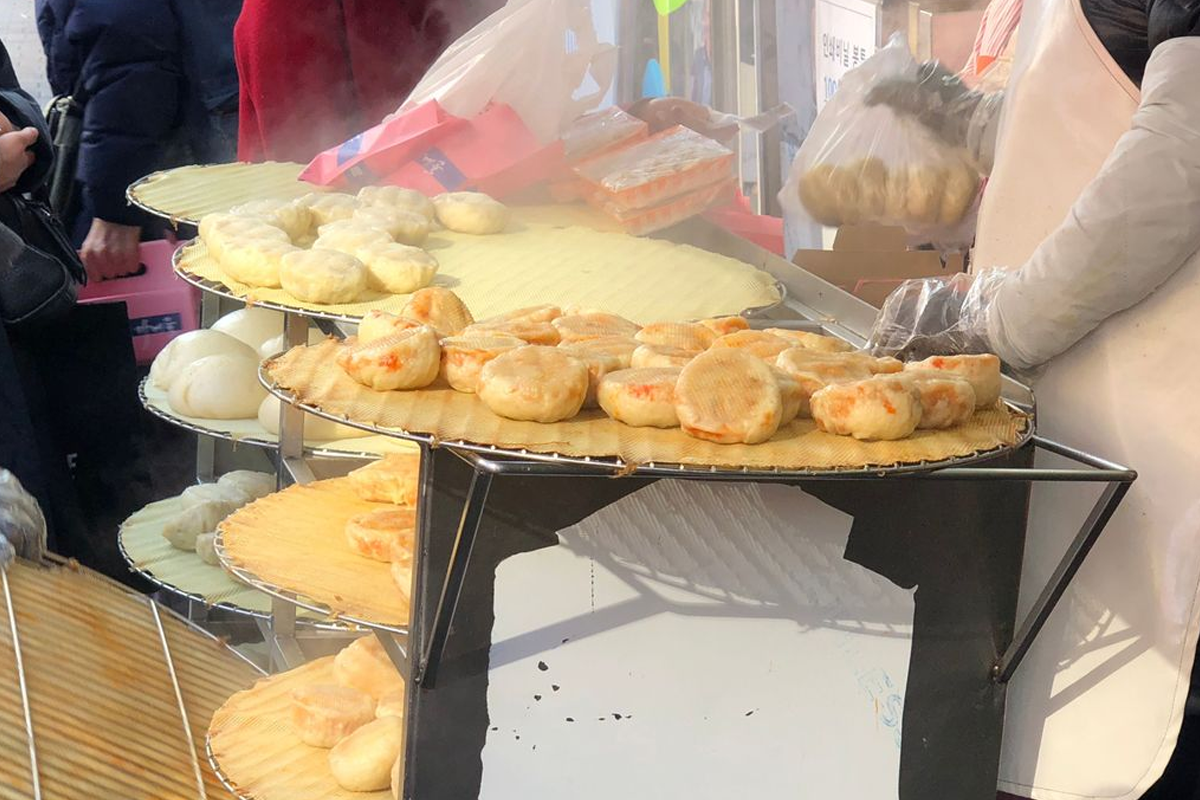 南大門市場裡的食物攤位，展示著多層陳列的韓式糖餅，蒸汽從熱騰騰的食物上升起，吸引旅客駐足品嚐。