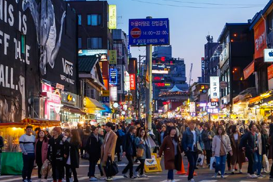 人群在夜晚行走於首爾弘大的一條繁華街道上，兩側有許多發光的商店招牌和光鮮亮麗的燈光，營造出活力四溢的城市氛圍。