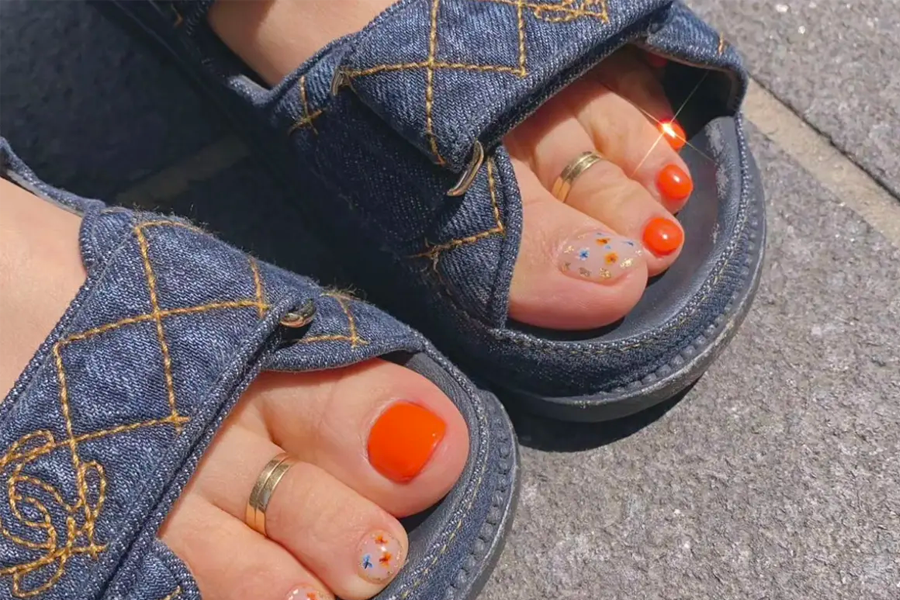 穿著涼鞋的腳部美甲，展示鮮橙色和花卉圖案的鮮明腳部美甲設計。