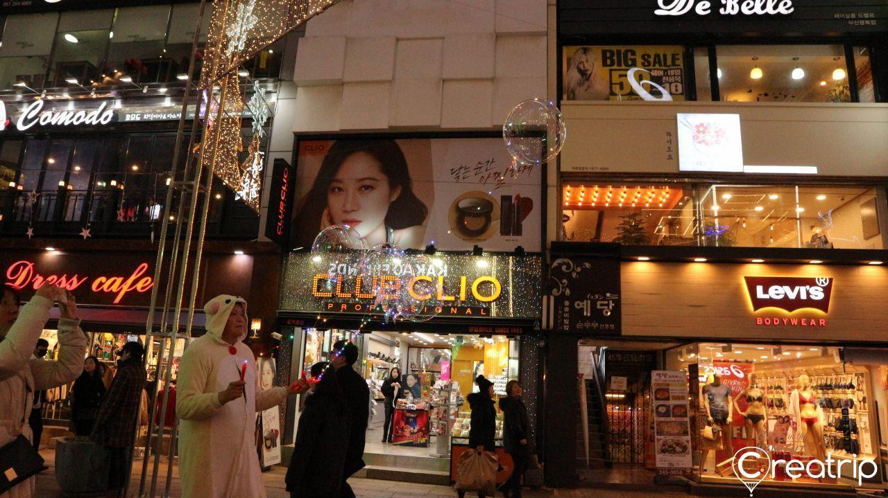 釜山繁华商业街夜景，商店霓虹灯标识闪烁，行人在街头来往。