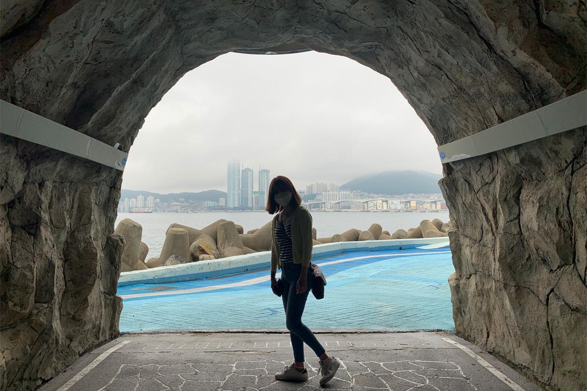 Woman posing at the entrance to the Huinnyeoul coastal Tunnel with the ocean and city in the background.