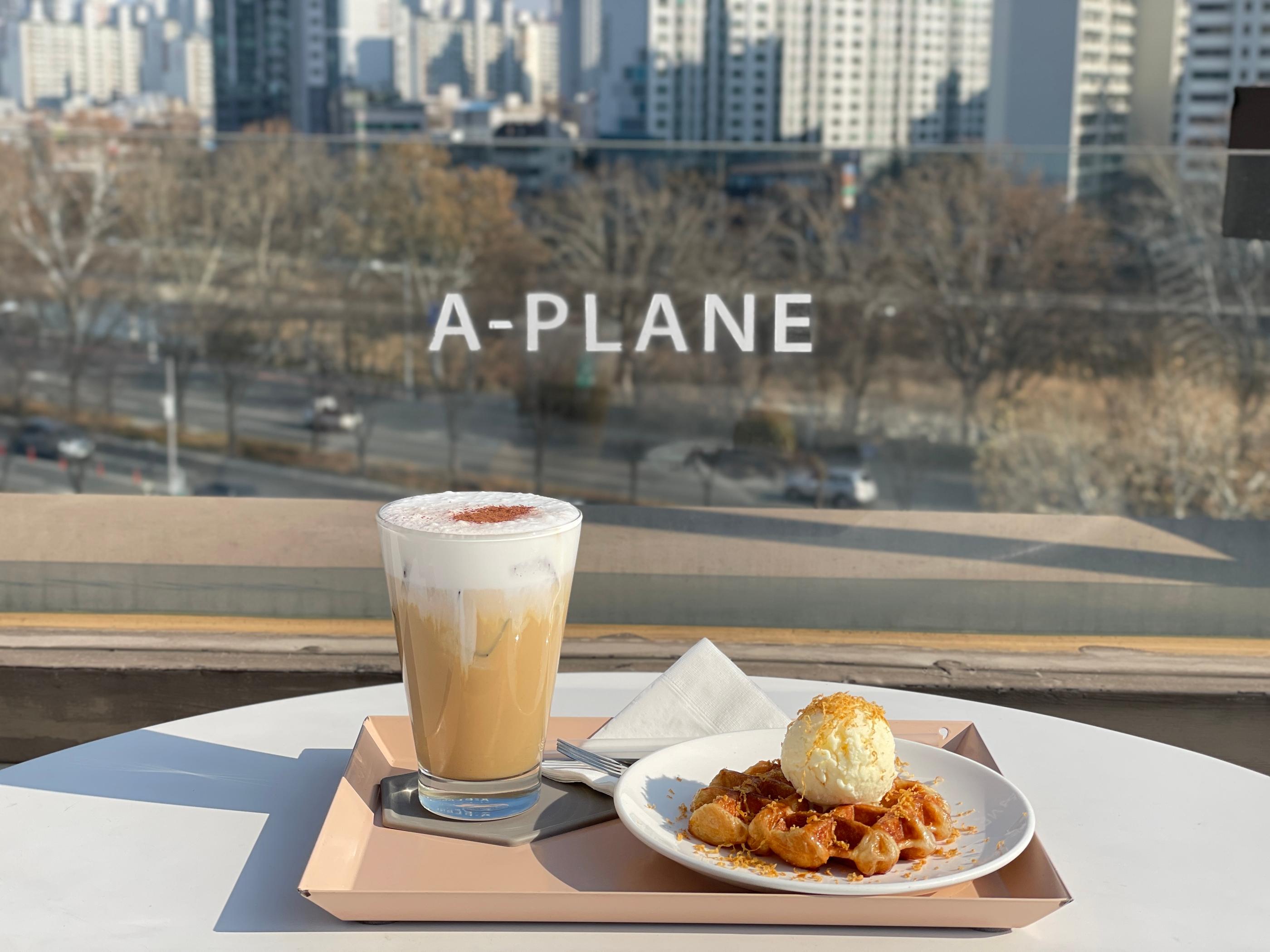 エイプレインで提供されるクリームラテとブラウンチーズクロッフル、カフェの眺望を背景にテーブル上に美しく配置されている