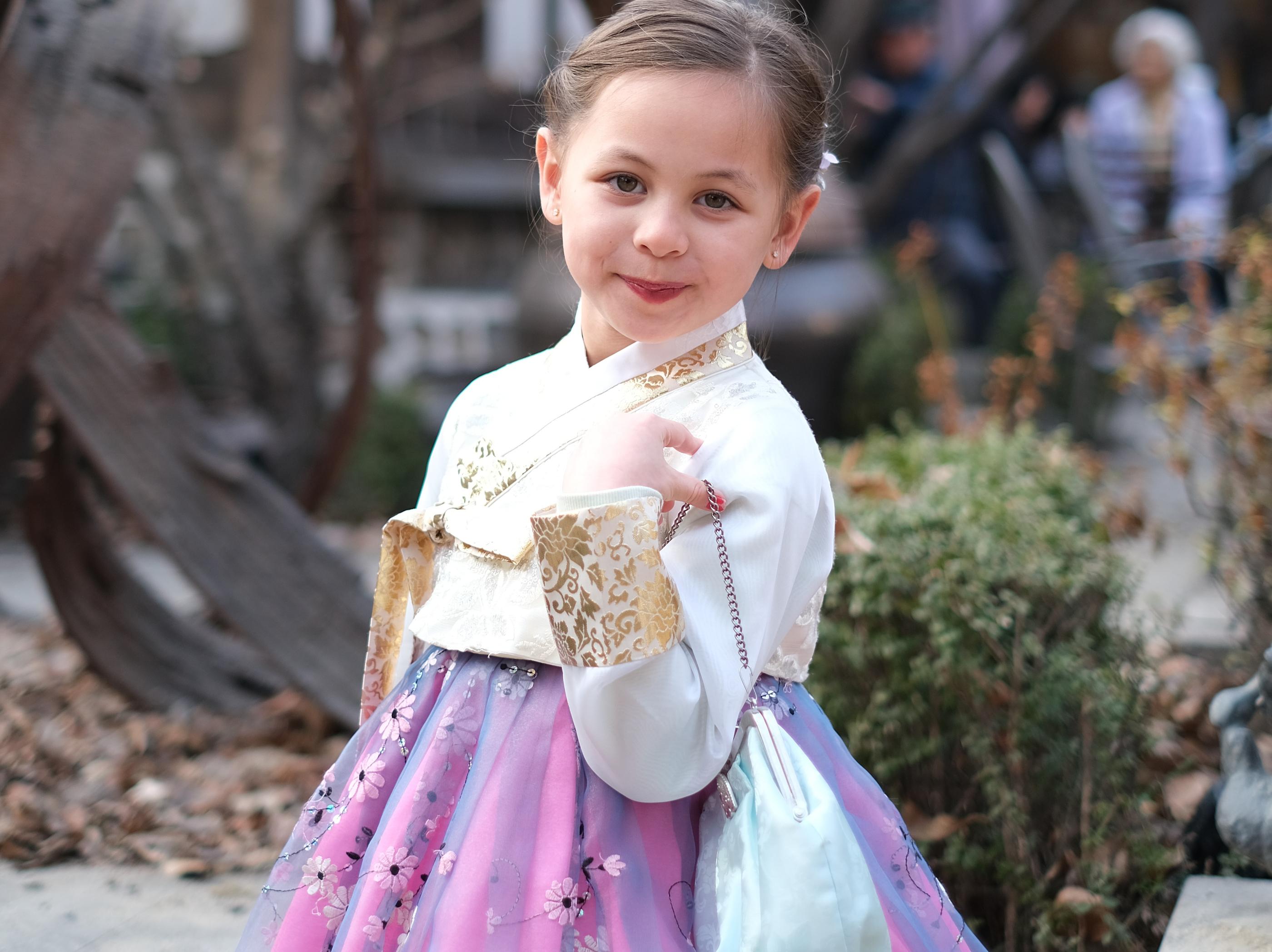 一名小女孩穿著韓服，手提銀色小袋子，背景是充滿韓國傳統氛圍的庭院。