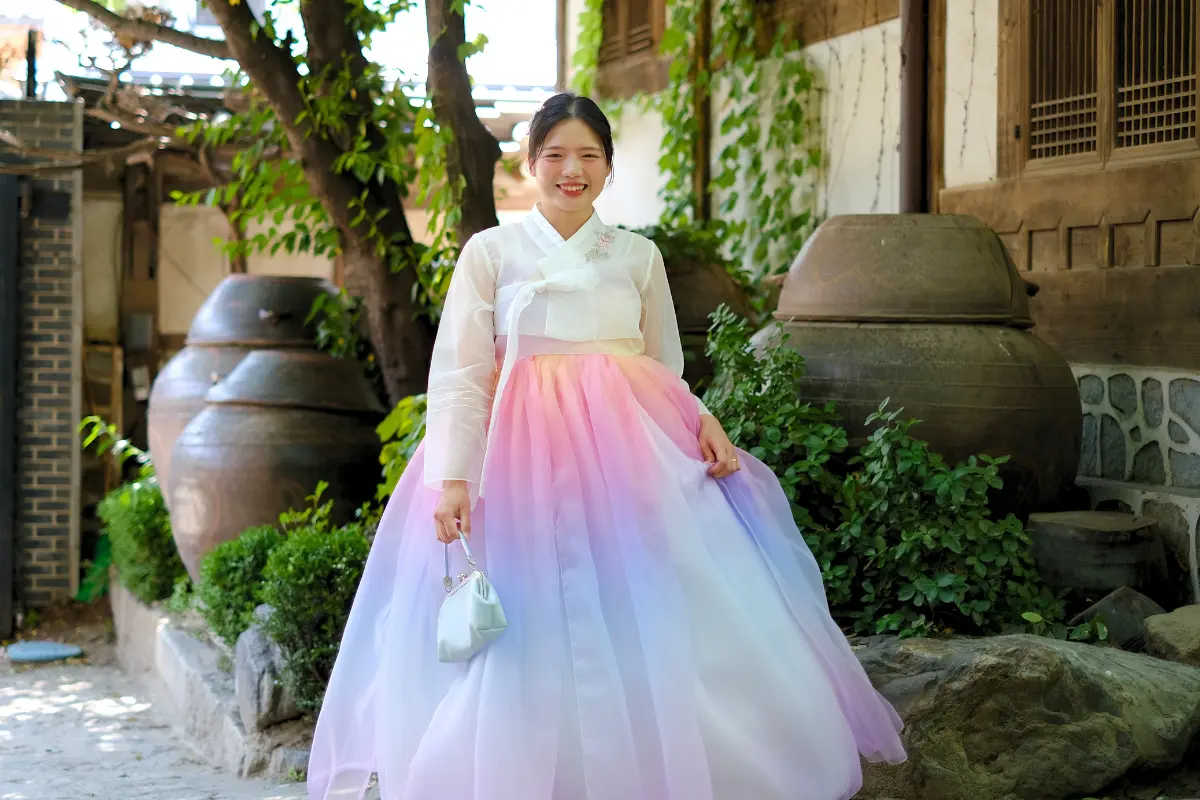 一位女子身穿粉色和彩虹漸變韓服，韓國傳統韓屋背景襯托出服飾的華美。