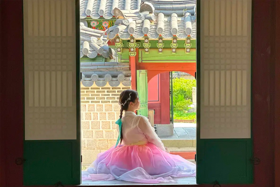 Entrance to a hanok village in Seoul, with a woman in hanbok seated under a traditional roof, creating an authentic cultural atmosphere.