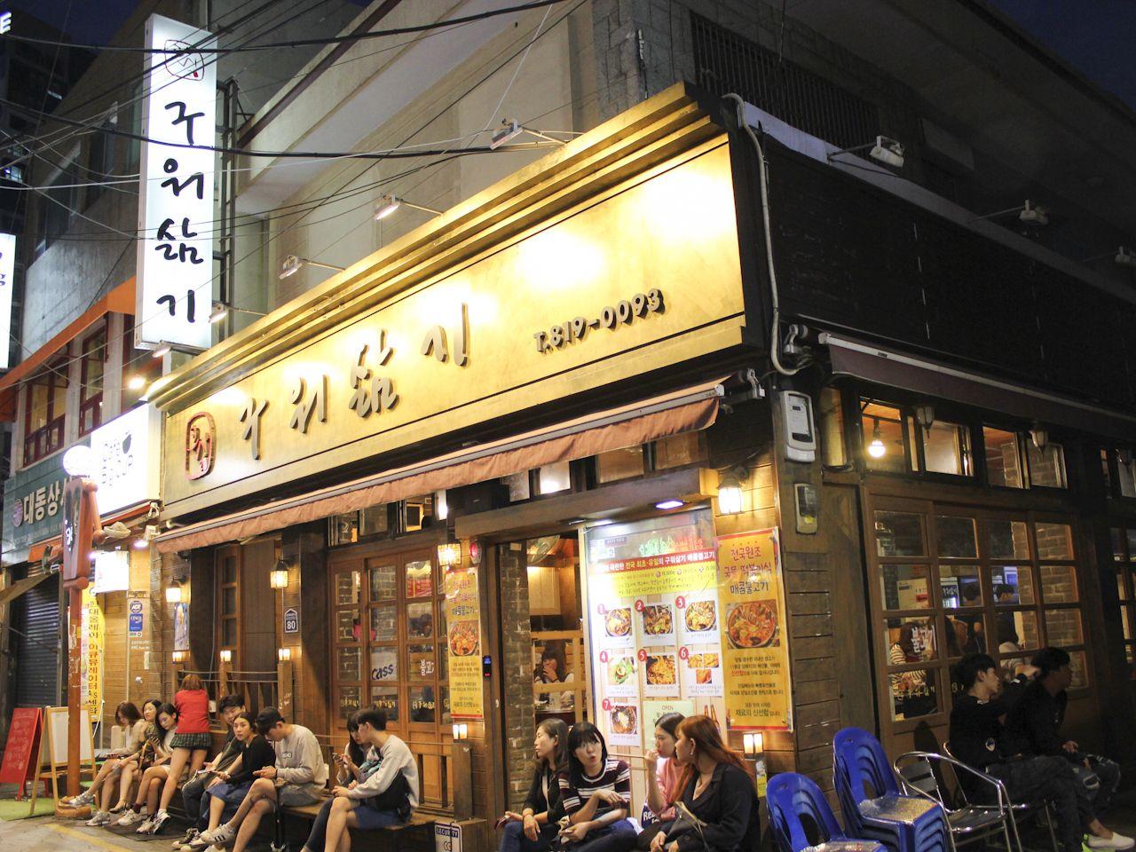 釜山“烤了又煮”餐廳，夜晚亮燈，窗戶透出暖黃的燈光， 外面坐著在等位的顧客