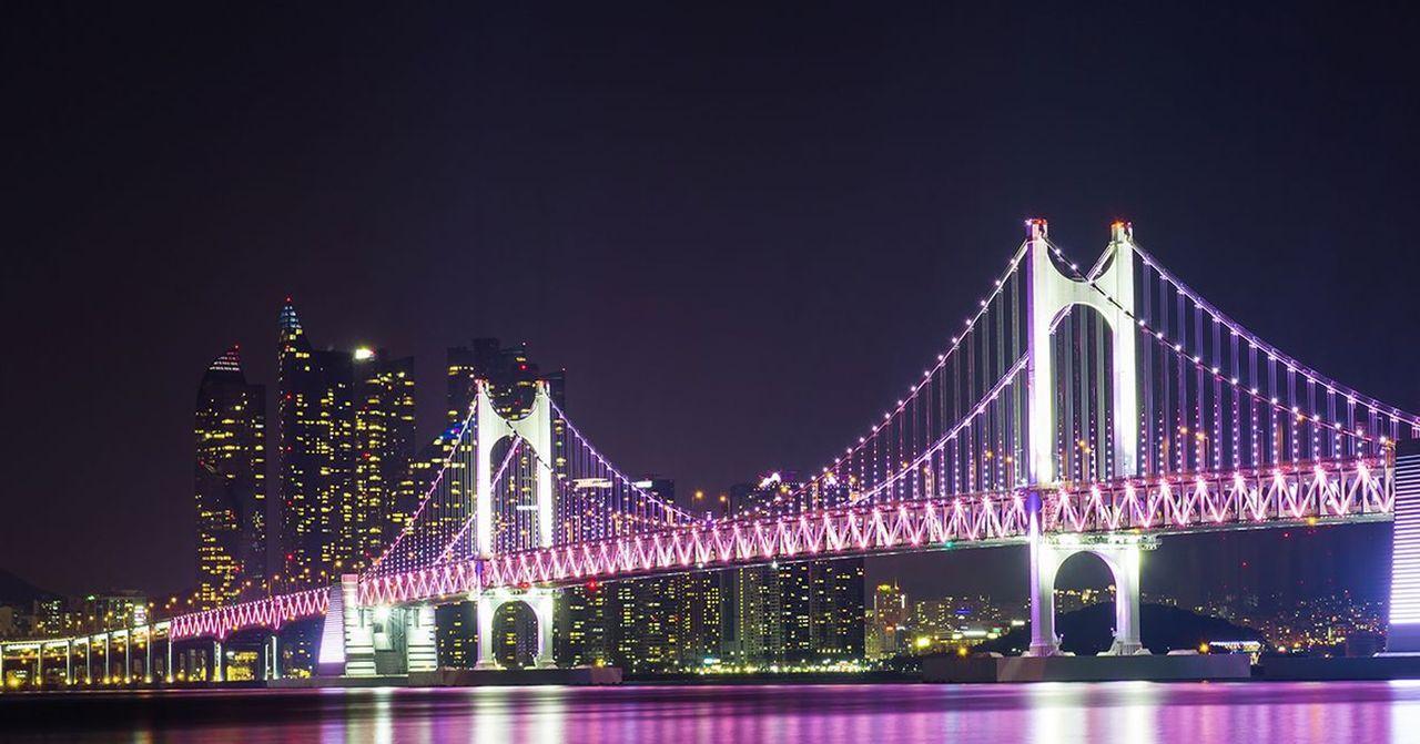 釜山夜景中璀璨的廣安大橋，橋身被燈光裝飾，背景是現代摩天大樓的天際線。