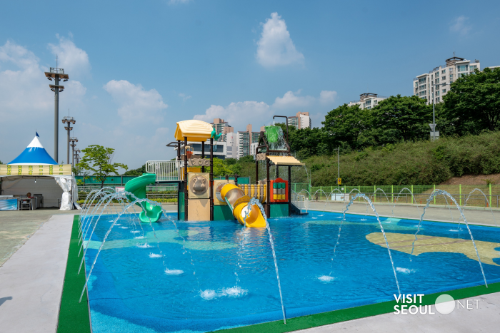 漢江公園兒童戲水區全景：藍天白雲下的淺水池、彩色滑水道與噴泉，適合家庭避暑與親子同遊，旁有帳篷與遠處高樓作為首爾場景
