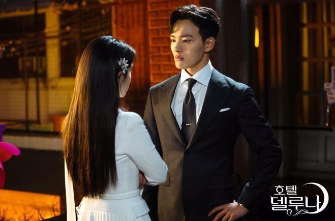 Deux personnages de 'Hotel Del Luna' se font face lors d'une scène tourmentée, capturant un moment de tension sur fond de rues animées.