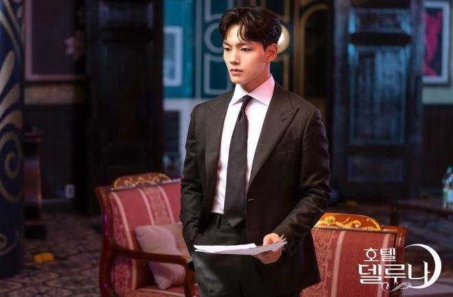 Un homme élégant dans un décor luxueux de 'Hotel Del Luna', portant un costume noir et tenant des papiers.