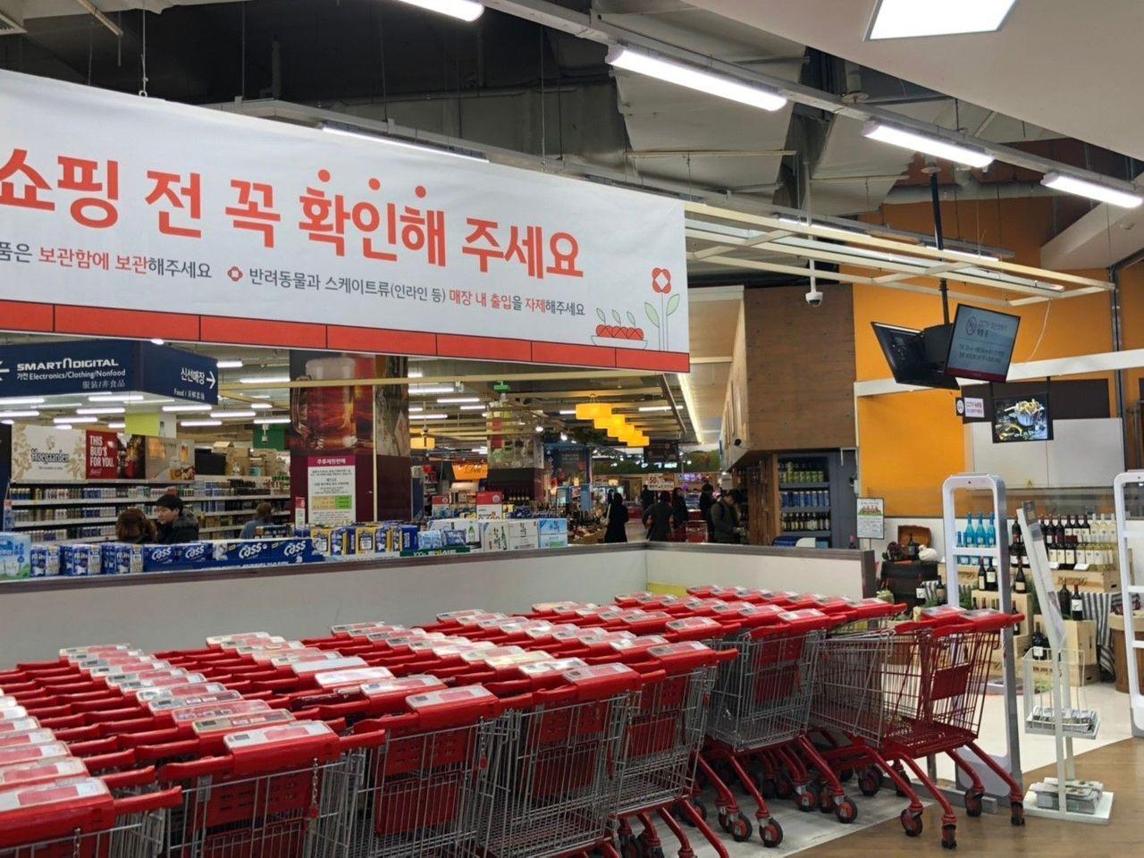 韩国超市内部，购物车排成一列，横幅标语显示购物前确认提醒。