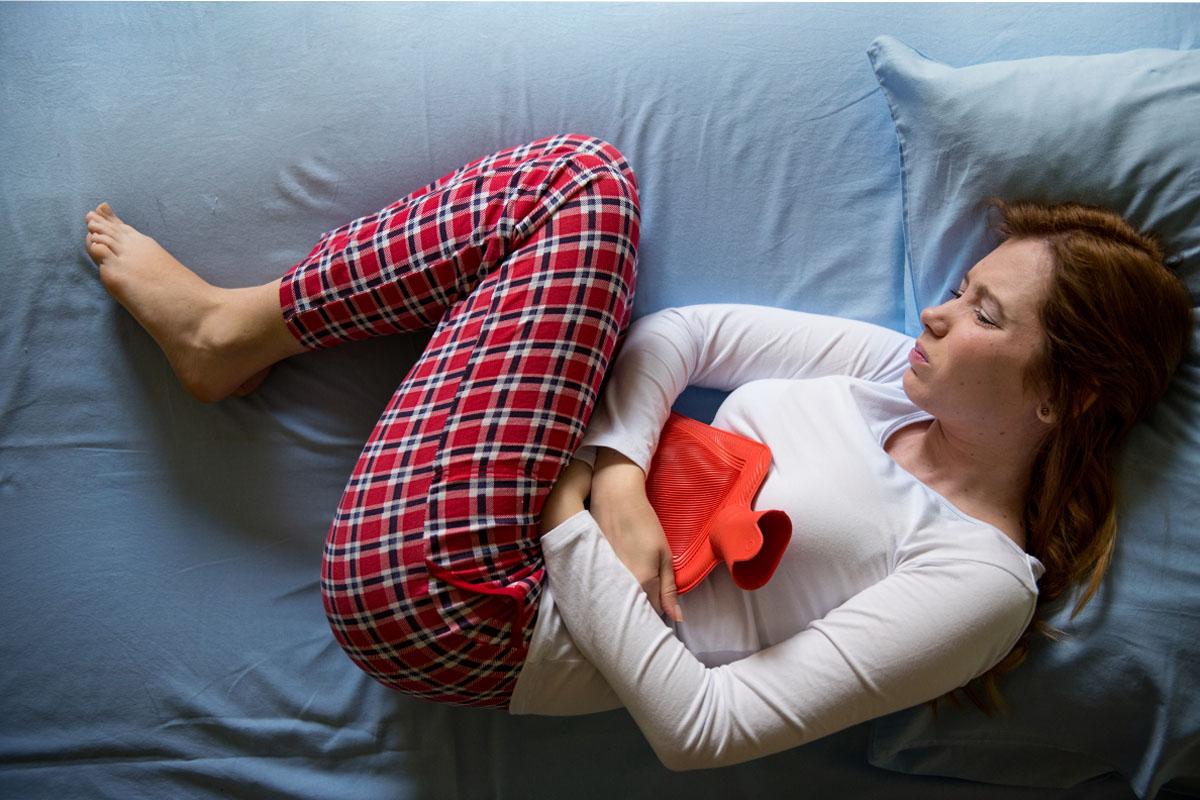 Hình ảnh một người nằm trên giường, ôm một chai nước ấm vào bụng trong tư thế cuộn tròn, biểu thị triệu chứng đau bụng kinh được đề cập trong bài viết về từ vựng tiếng Hàn.