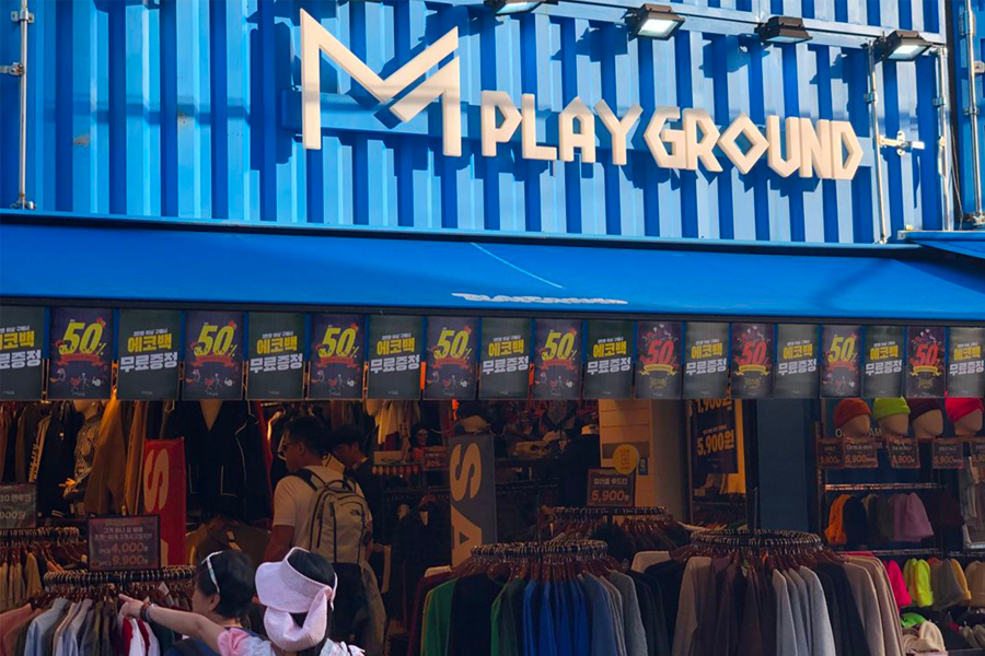 M PlayGround弘大店的外觀，顯示店面的藍色建築和服裝價格。」