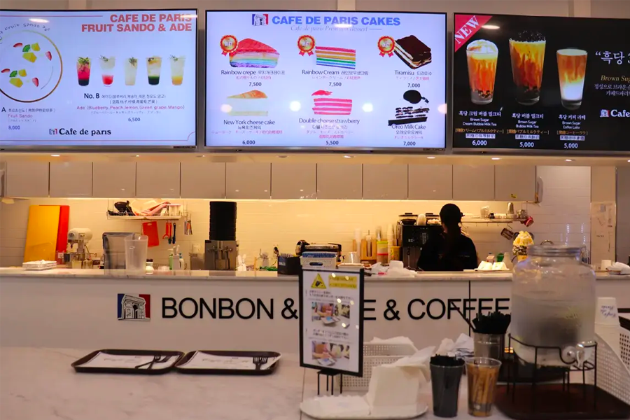 카페 드 파리의 주방 카운터로, 메뉴와 가격 표시가 있는 전광판이 있는 모습입니다.