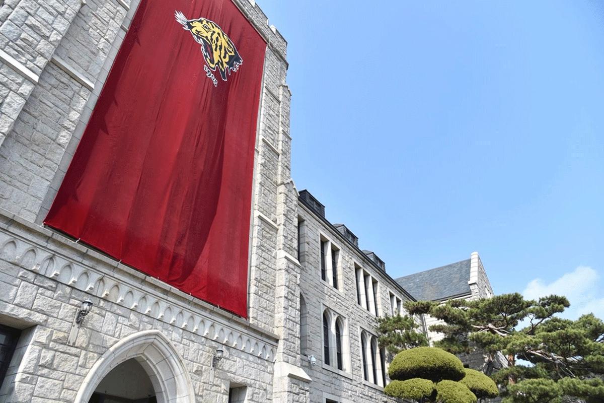 Außenansicht der Korea University mit einem großen roten Banner mit dem Logo des Tigers, das an der Fassade eines steinernen Gebäudes hängt. Die Fassade zeigt gotische Architekturelemente und ist von einem klaren blauen Himmel umgeben, was die beeindruckende und prestigeträchtige Atmosphäre dieser renommierten Bildungseinrichtung unterstreicht.
