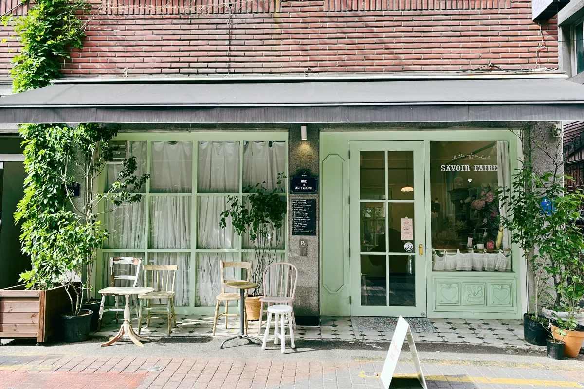 Exterior of a stylish Korean café with green door and window frames, welcoming with chairs and plants outside.