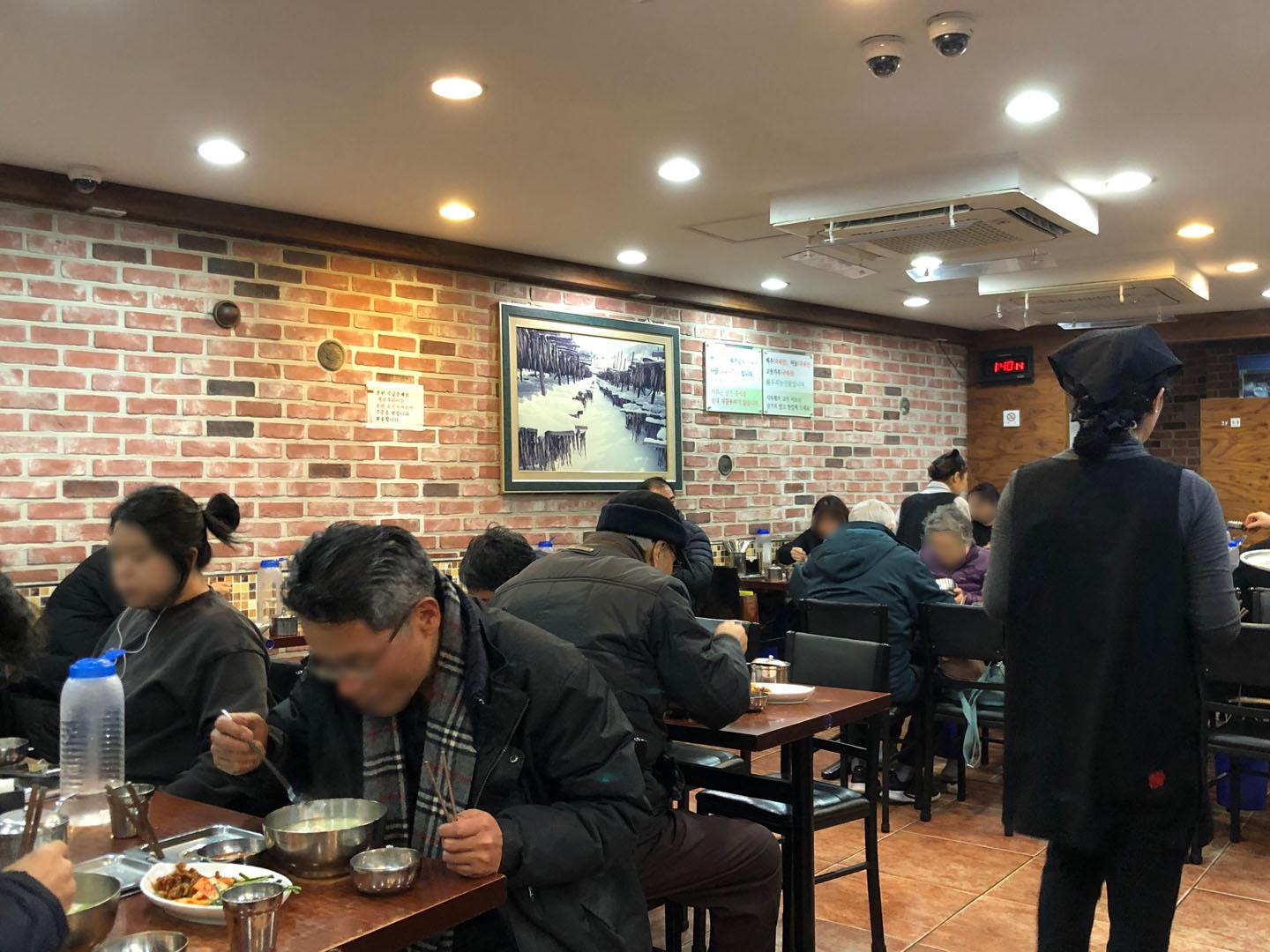 ウルチロ入口駅付近にある武橋洞プゴグッチッの店内風景。多くの客が食事を楽しんでいる様子。テーブルには幹し鱈スープと豊富な付け合わせが並び、活気に溢れている。