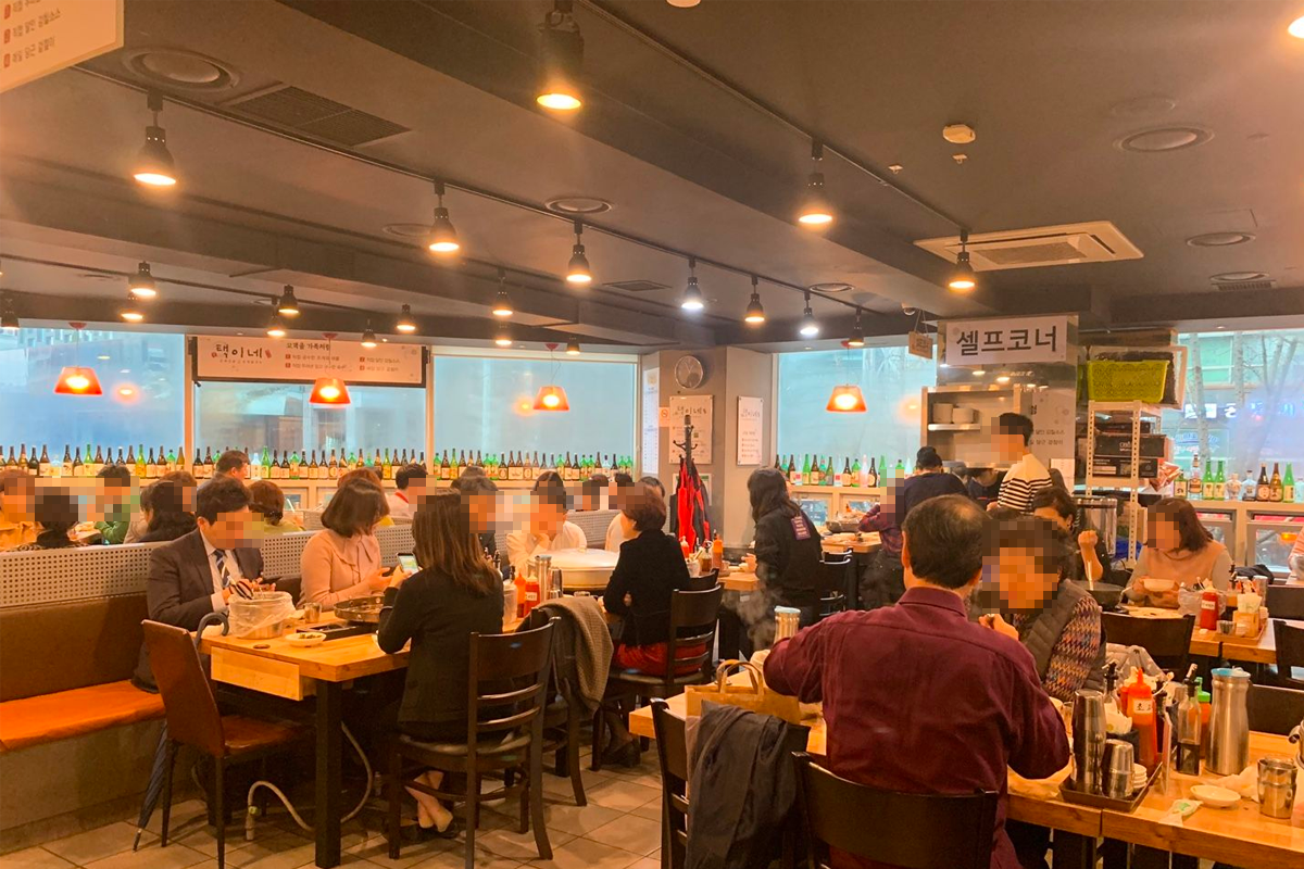 韩国钟路区的Jogae Jeongol餐厅的内景，展示了顾客在享用美食。店内装饰温馨，背景可见韩文菜单和装饰灯。