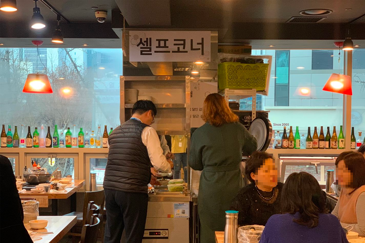 在泽家贝壳蒸锅餐厅的自助区，顾客正在自助取食，背景墙上排列着韩国烧酒瓶，餐厅氛围热闹而现代。