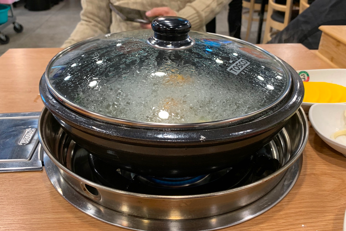 一份清淡的白湯內包含新鮮牛肉片、各類蔬菜及手工餃子，旁邊配有韓式泡菜和小菜。