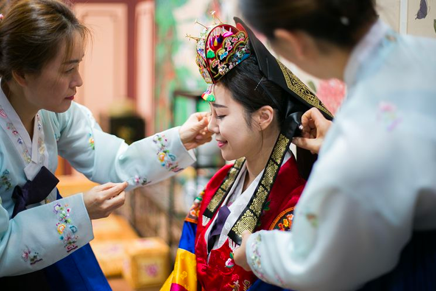 兩位穿著色彩鮮豔韓服的女性幫戴傳統婚禮頭飾的女性整理服裝，背景是室內場景。