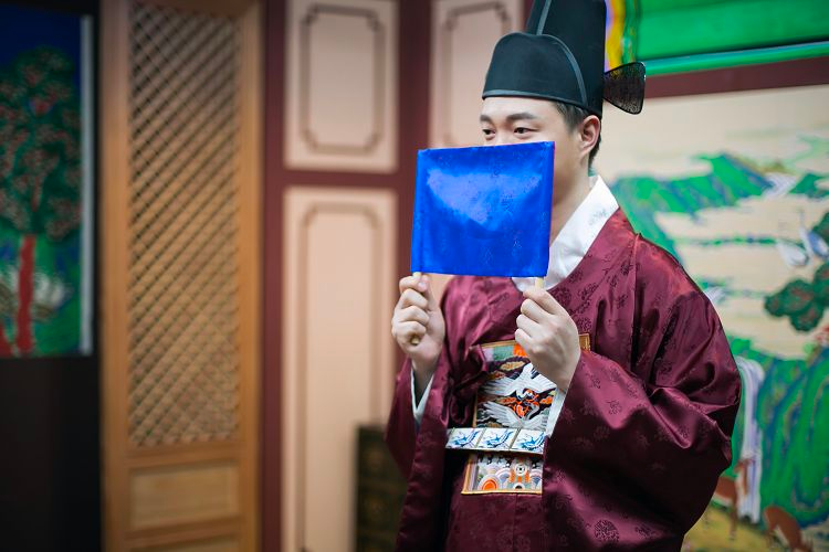 身穿紅色韓服的男性站在室內場景中，手持藍色扇子，背景有韓國傳統風格的牆壁裝飾。