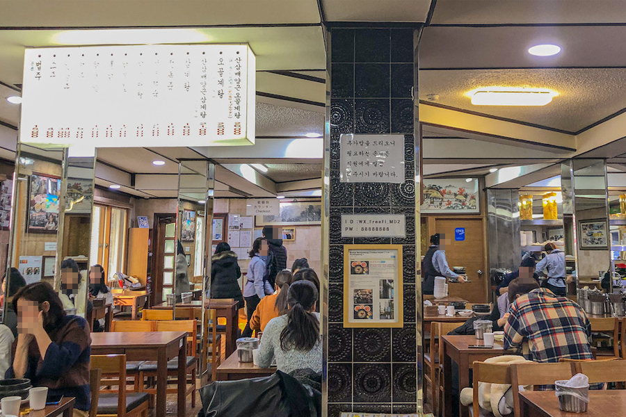 Innenaufnahme des Baekje Samgyetang Restaurants in Myeongdong mit Kunden, die ihre Mahlzeiten genießen. Traditionelle Tischanordnung und Dekoration, die eine gemütliche Atmosphäre schaffen.