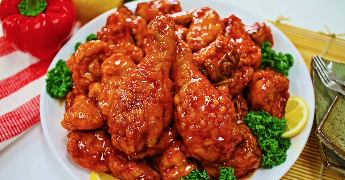 一大盘韩国炸鸡，上面覆盖着浓郁的甜辣酱，围绕着柠檬片和绿色蔬菜装饰。