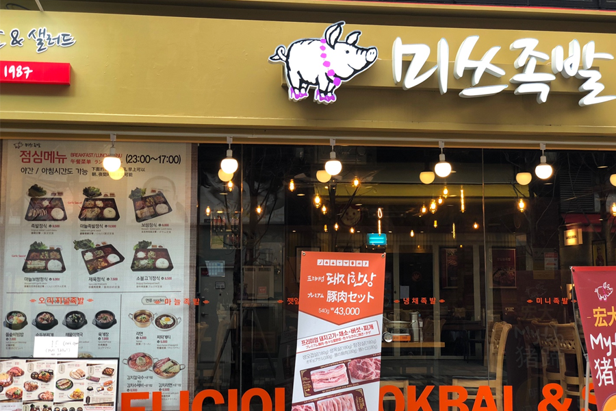 位於首爾明洞的著名餐廳「豬腳小姐」外觀，餐廳招牌上有豬腳的圖案，店門口展示著特別菜單的海報。