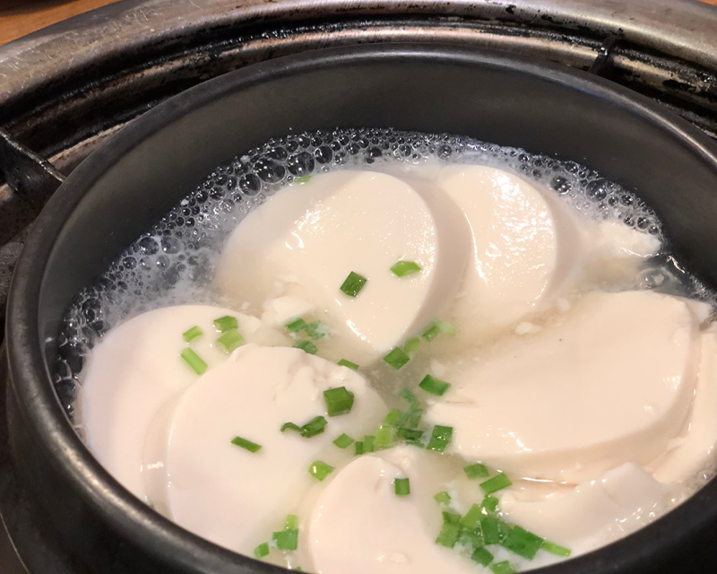 一鍋新鮮白豆腐在滾燙的湯中煮沸，撒上葱花增添香氣，適合搭配豐富的烤腸料理。