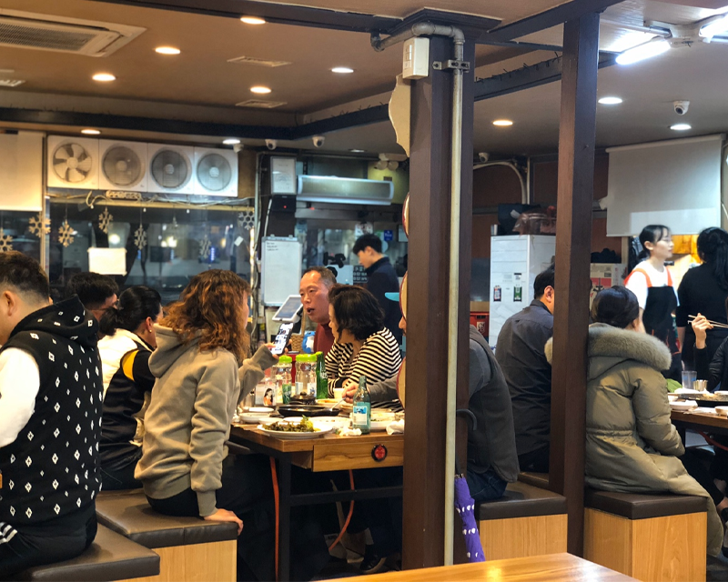 店內熙熙攘攘，食客們在緊湊的桌椅上享用韓式烤腸，熱鬧非凡的用餐氛圍。