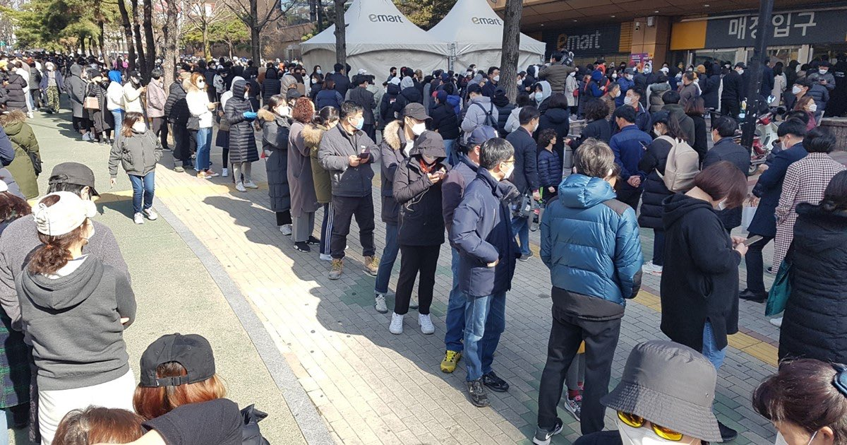 一羣戴著口罩和冬季衣物的民眾在户外排隊，等待進入韓國的大型零售商店，顯示疫情下的口罩搶購潮。
