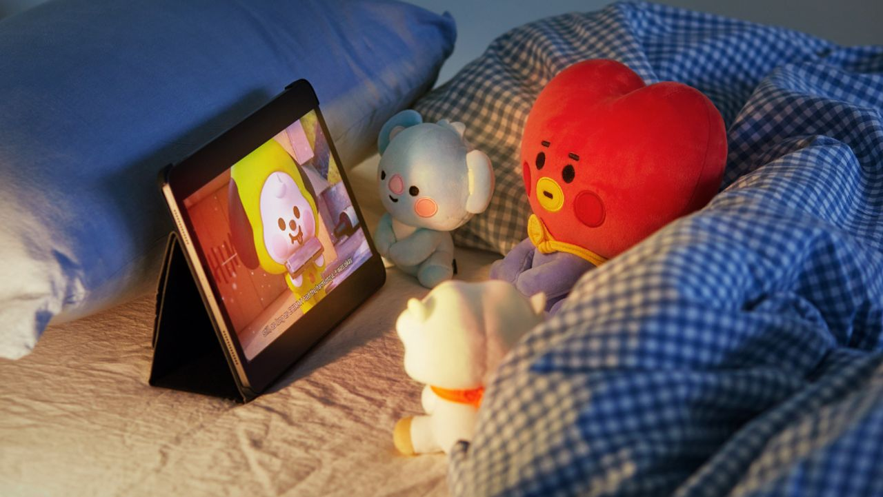 BT21 Baby玩偶观看平板电脑上的动画，玩偶分别是经典的BT21角色如TATA和CHIMMY，这张舒适的床头场景中营造出温馨的氛围。