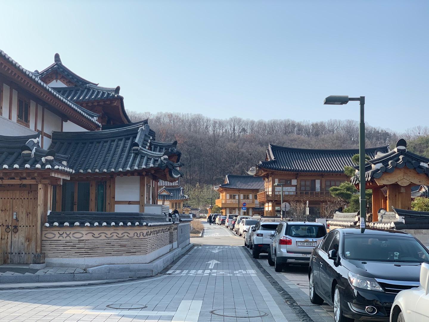 Street view of Eunpyeong Hanok Village with parked vehicles along the road, traditional wooden Korean houses on both sides, and a forested area in the distance.