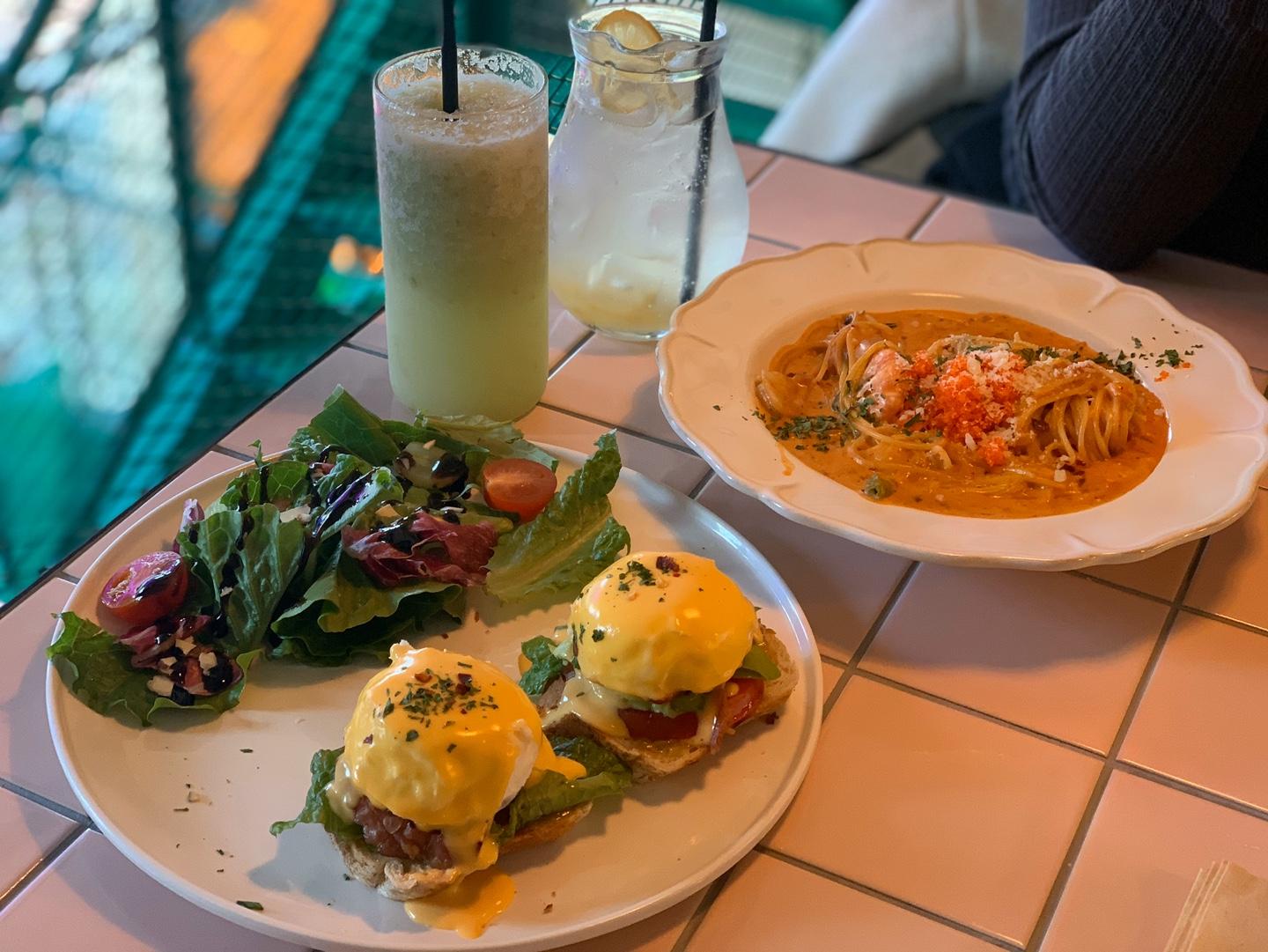 Table at a cafe with brunch dishes including Eggs Benedict, shrimp rosé pasta, and fresh salads on a white tiled tabletop.
