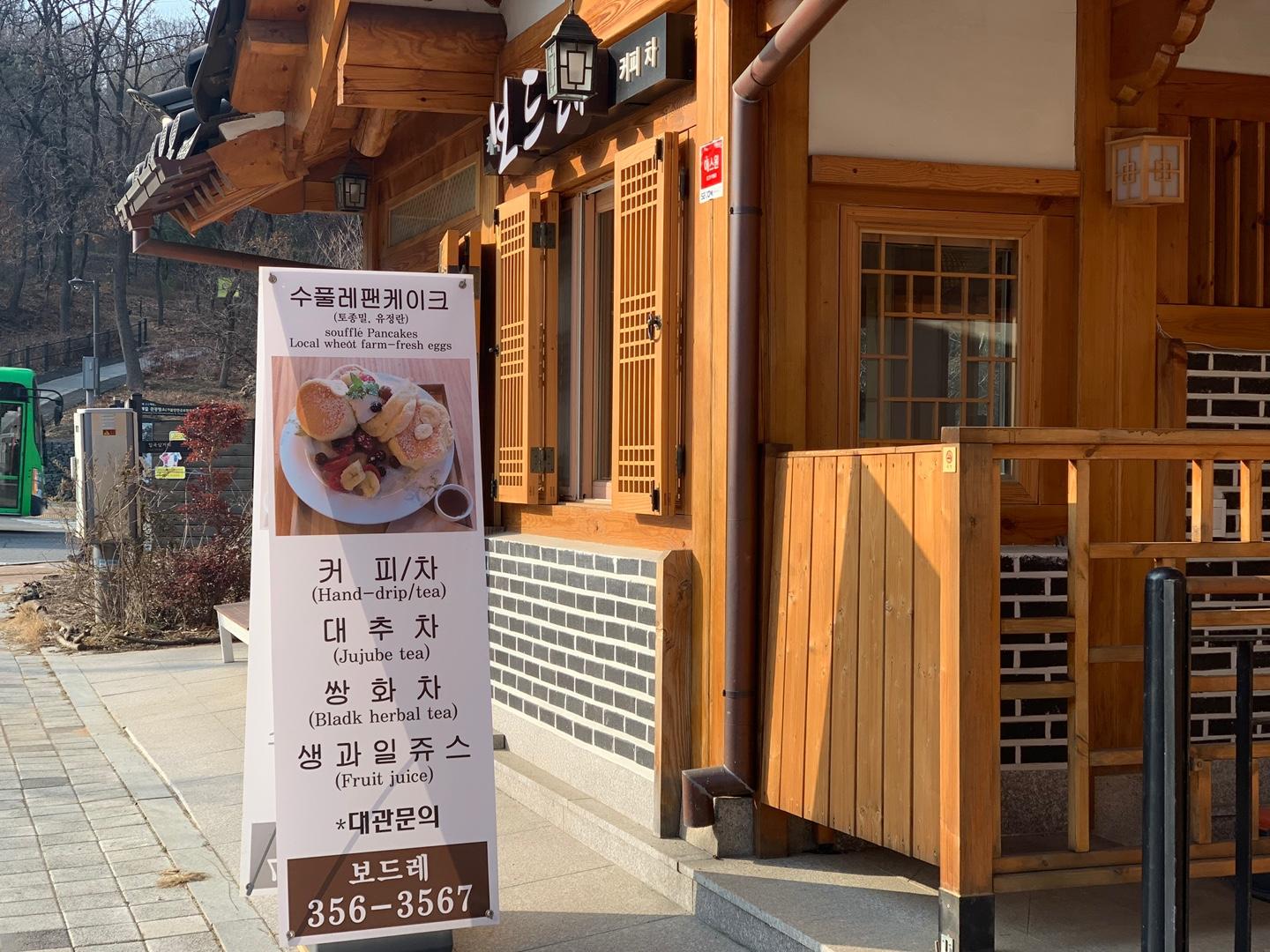 Sign outside a hanok cafe in Eunpyeong Hanok Village advertising souffle pancakes and various teas with the wooden architecture of the cafe visible in the background.