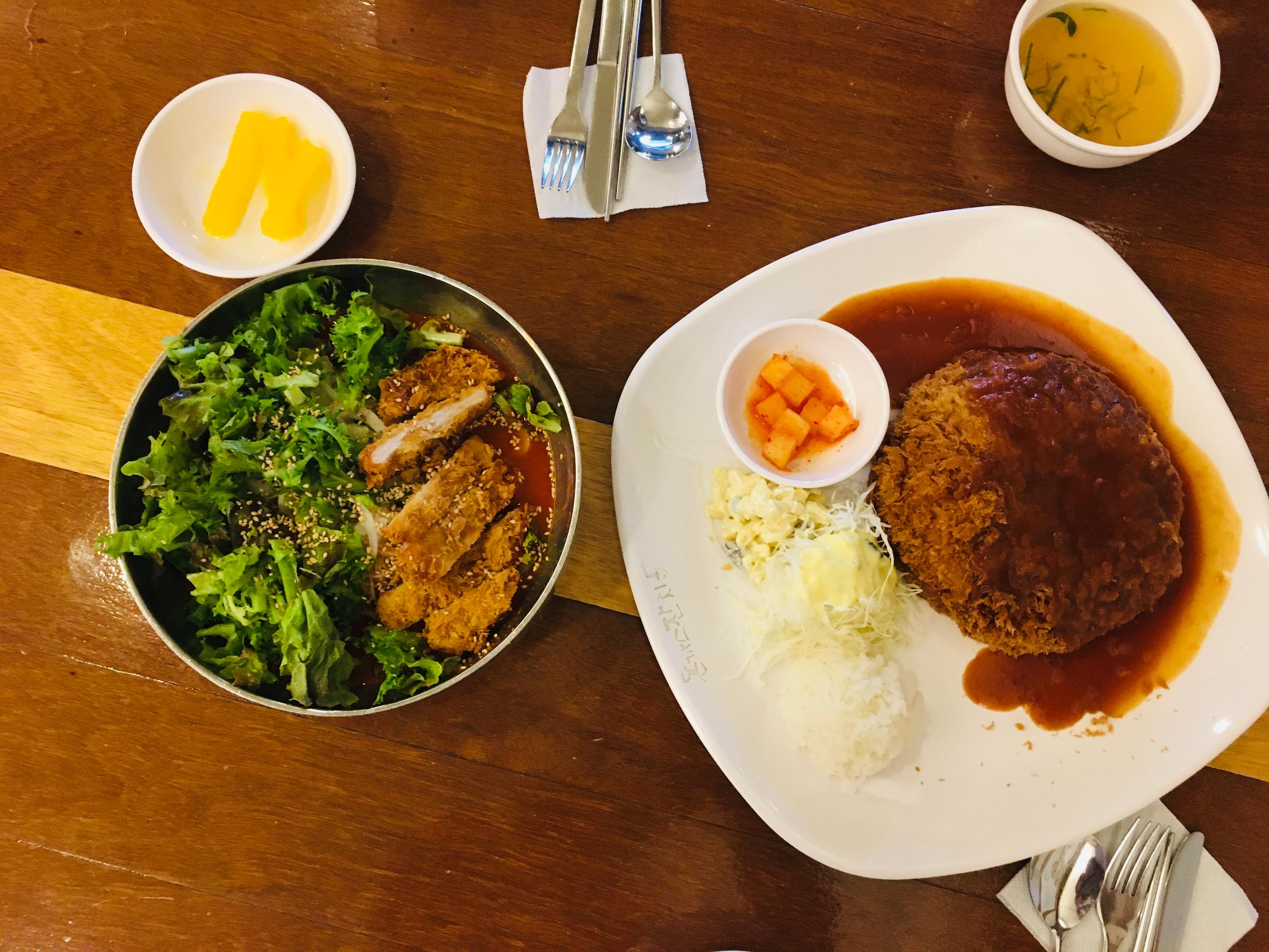 An overhead view of a delicious Korean dinner set, featuring spicy noodles with crispy pork cutlets and a side of rice with salad and pickles, served on a wooden table at Dongkatsu Janchi restaurant in Itaewon, Seoul.