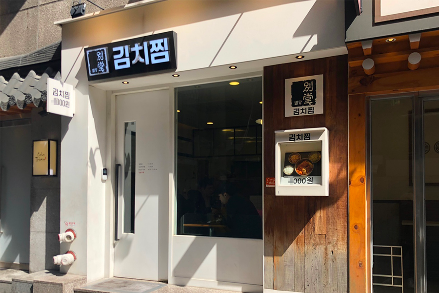 ป้ายร้านบยอลดัง กิมจิจิม ในชินชน กรุงเทพฯ แสดงการเข้าถึงที่ตั้งร้านอาหารอย่างง่ายจากสถานีชินชน พร้อมป้ายบอกสถานที่สลักคำว่า'กิมจิจิม'และมีรับแสงแดดอ่อนๆในเมือง