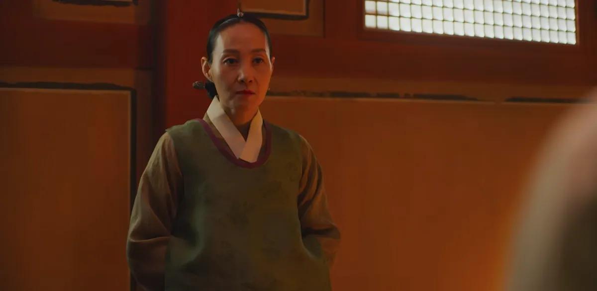 Escena de una mujer de pie en un entorno interior tradicional en 'Kingdom', con una vestimenta de sirvienta.